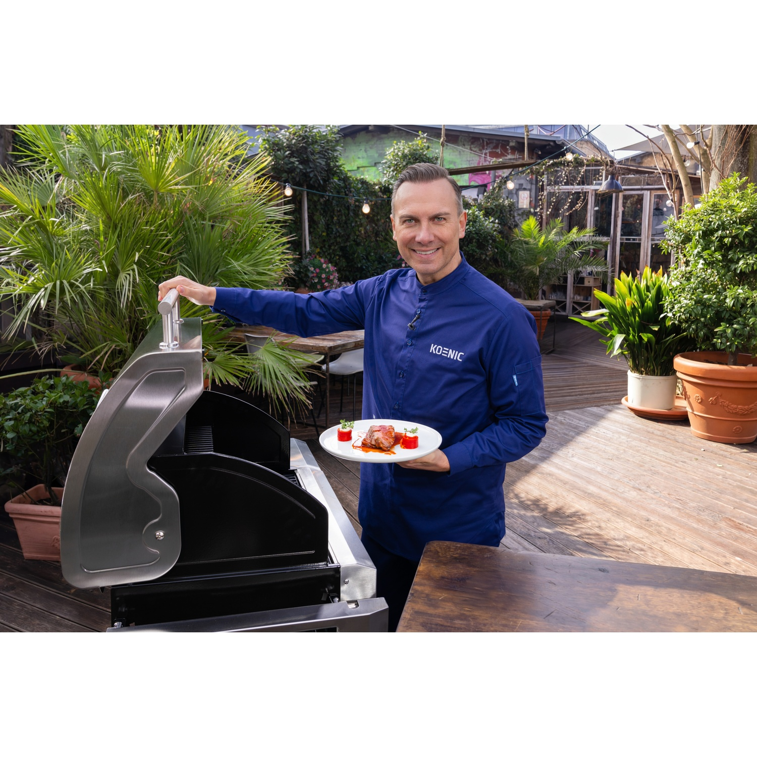 Homme en veste de chef bleue présente assiette de viande grillée et accompagnements près d'un barbecue.