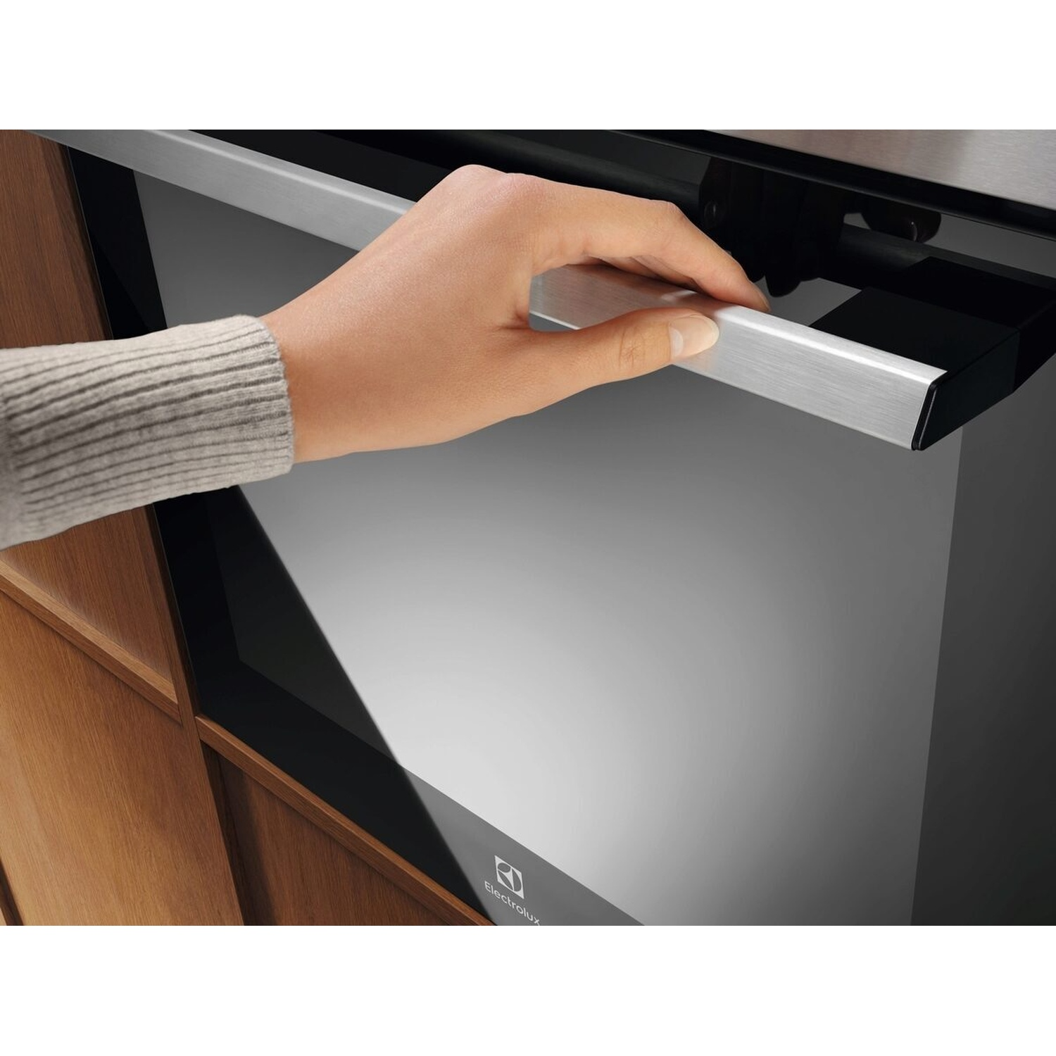 Una mano in un maglione grigio apre un forno moderno con maniglia in metallo spazzolato.