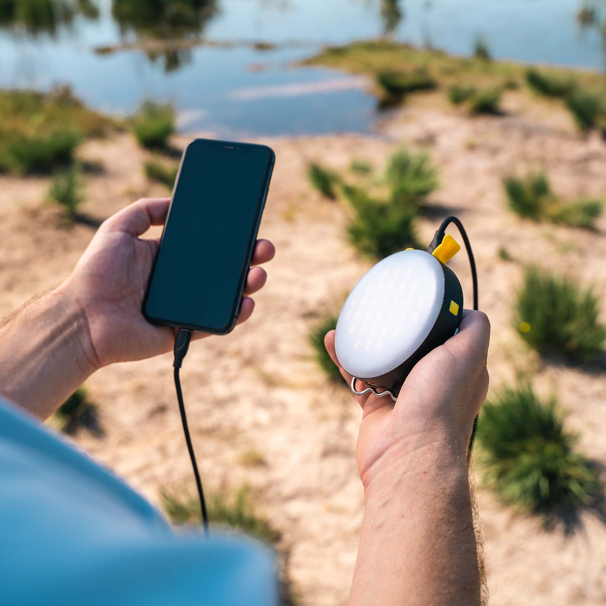 Eine Hand hält ein Smartphone, verbunden mit einem Ladekabel. Die andere Hand hält eine runde Lampe.
