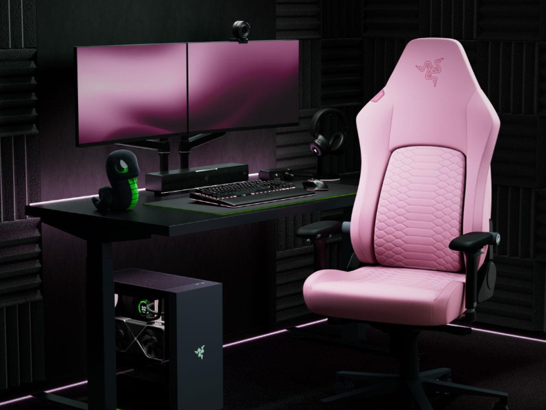 Różowe krzesło gamingowe obok biurka z dwoma monitorami i komputerem.