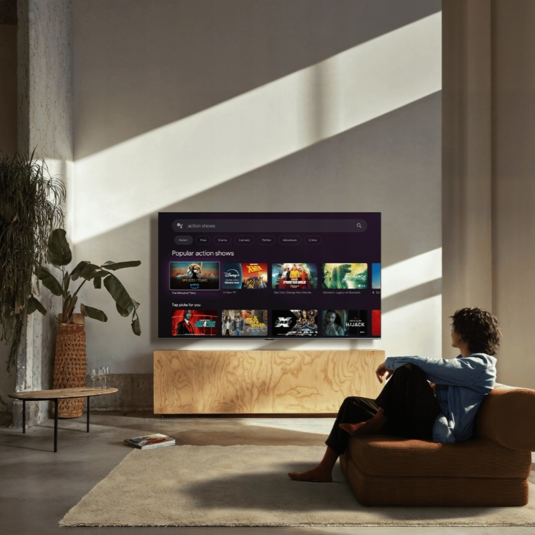 Osoba siedzi na brązowej kanapie i ogląda telewizor wyświetlający seriale akcji.