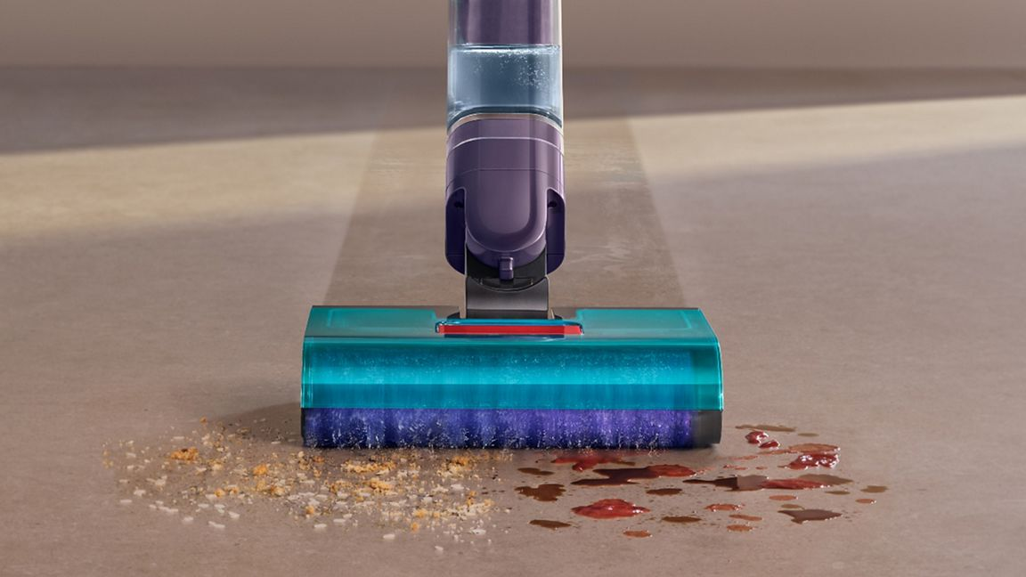 Un aspirateur violet nettoie des miettes et du liquide sur un sol.