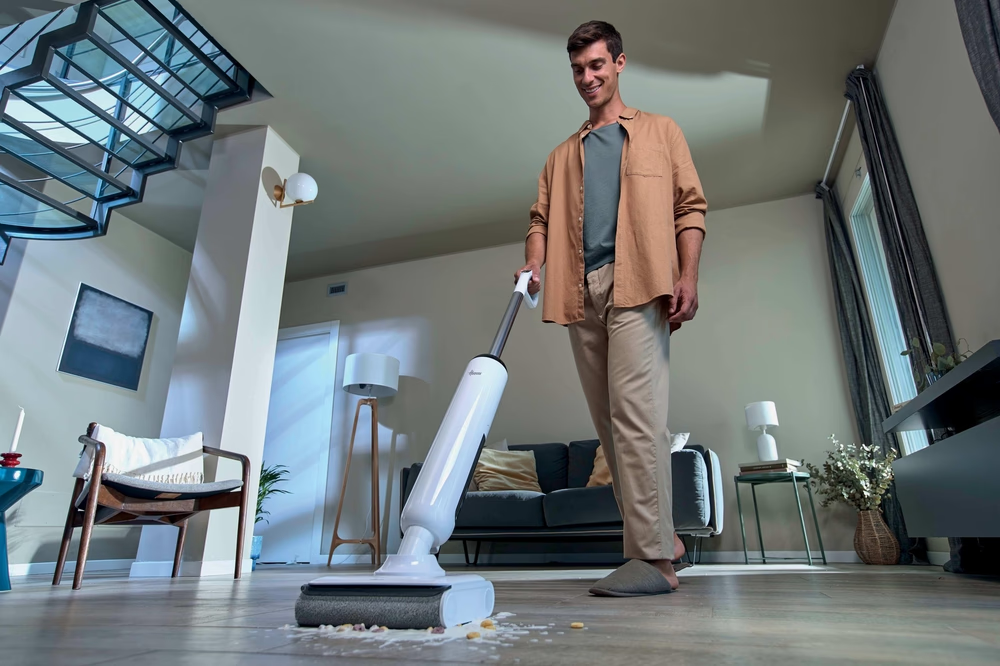 Mann saugt Essensreste mit einem weißen Bodenwischer in einem modernen Wohnzimmer auf.