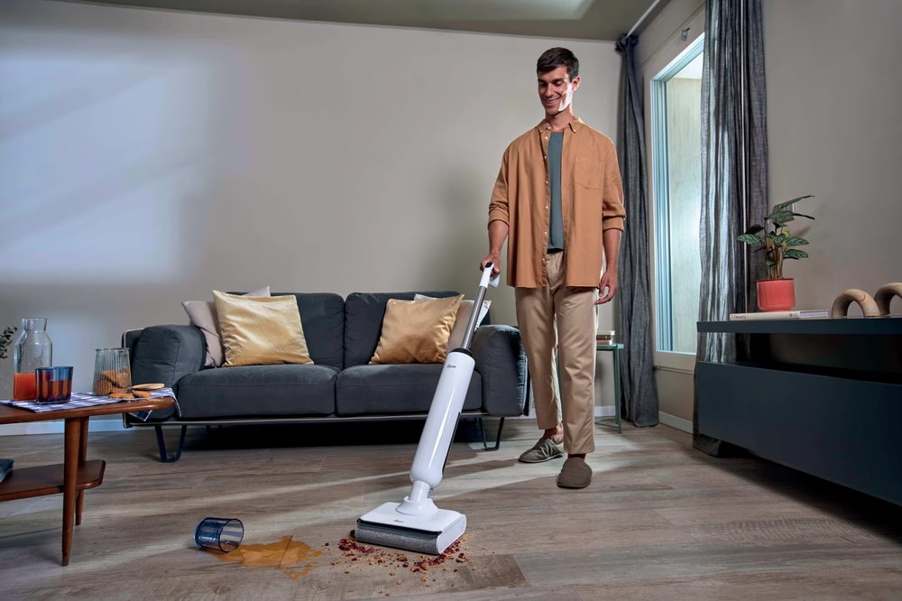 Mann reinigt verschüttete Flüssigkeit und Krümel mit einem Bodenreiniger in einem Wohnzimmer.