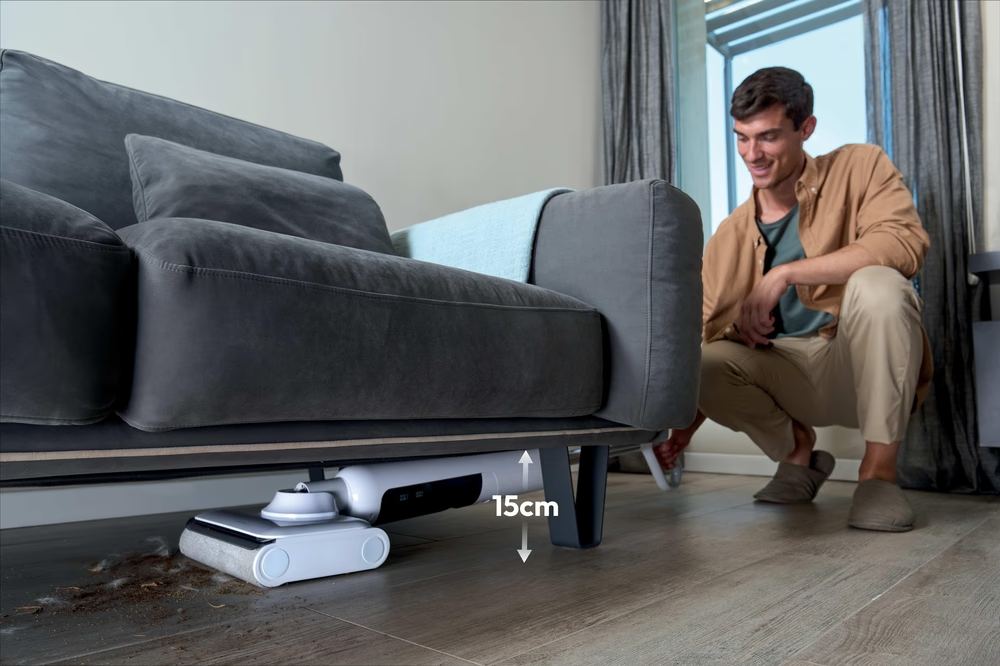 Ein Mann reinigt den Boden unter einem Sofa mit einem schlanken Staubsauger.