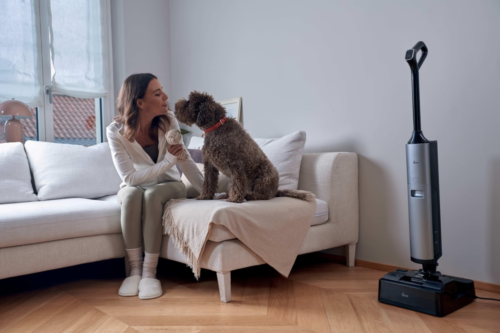 Eine Frau sitzt auf einem Sofa und spielt mit ihrem Hund. Ein Staubsauger steht daneben.