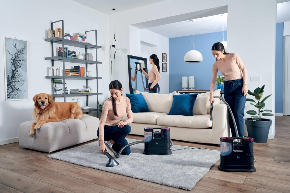 Zwei Frauen reinigen ein Wohnzimmer mit Staubsaugern, ein Hund liegt auf einem Hocker.