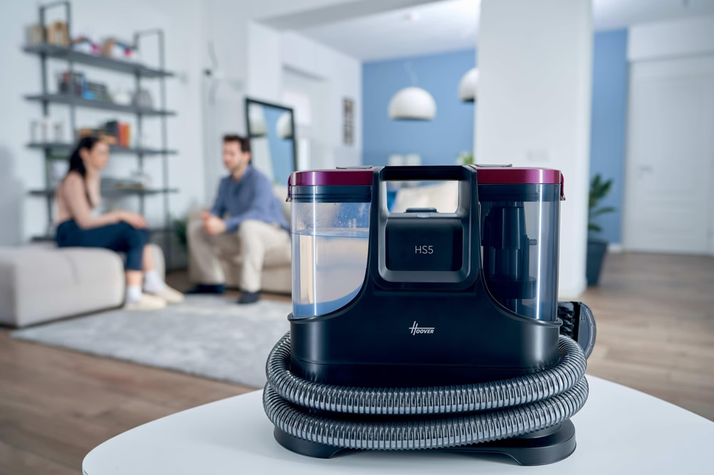 Dunkler Hoover HS5 Nass-/Trockensauger mit Schlauch auf Tisch. Paar im Hintergrund.