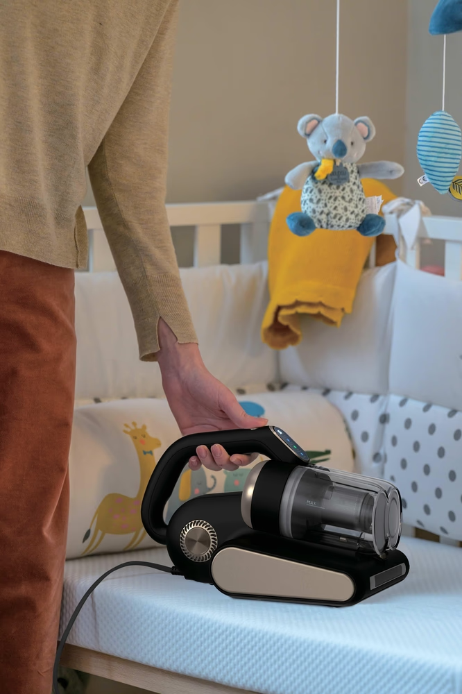 Eine Hand hält einen schwarzen Staubsauger über einem Babybett mit Spielzeug.