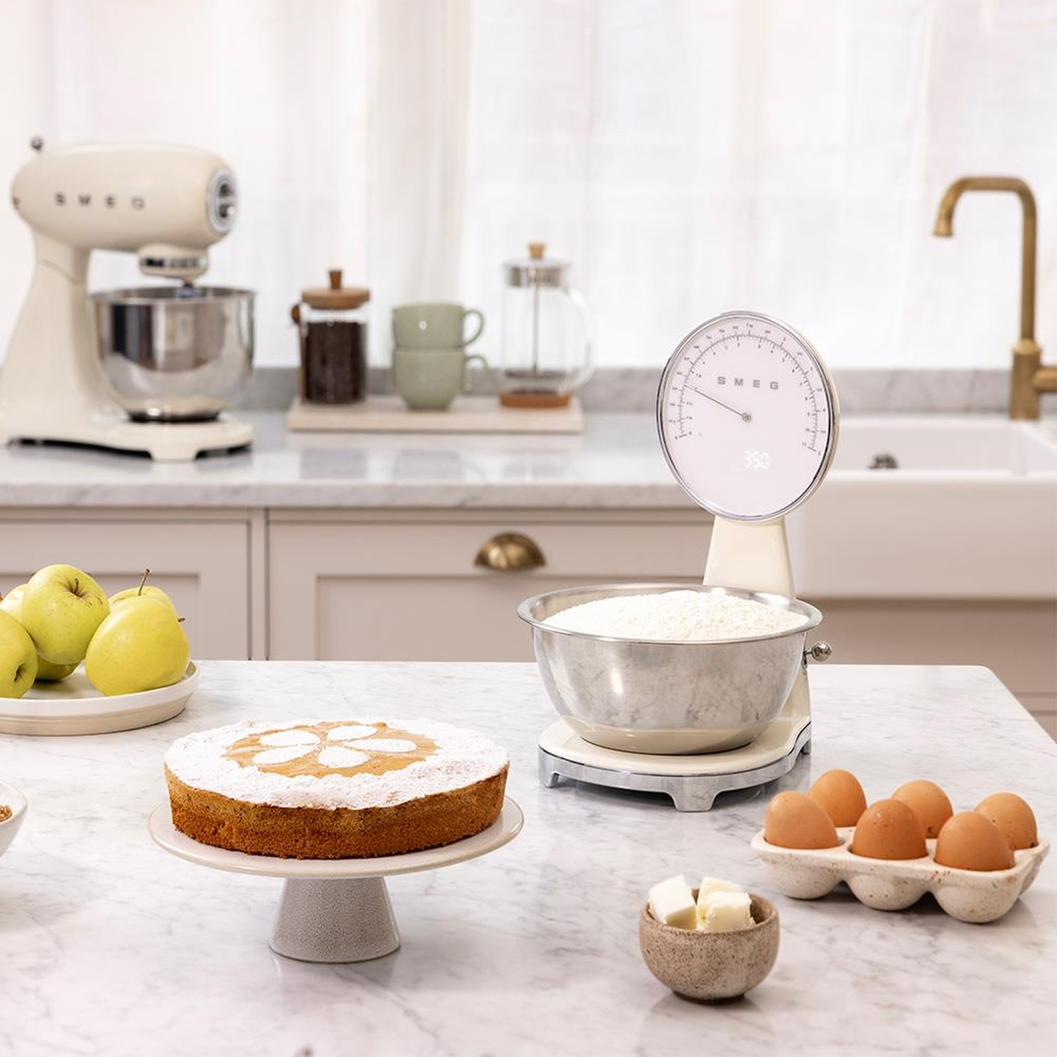 Una bilancia da cucina con farina, uova, torta, mele e impastatrice su un piano di lavoro in marmo.