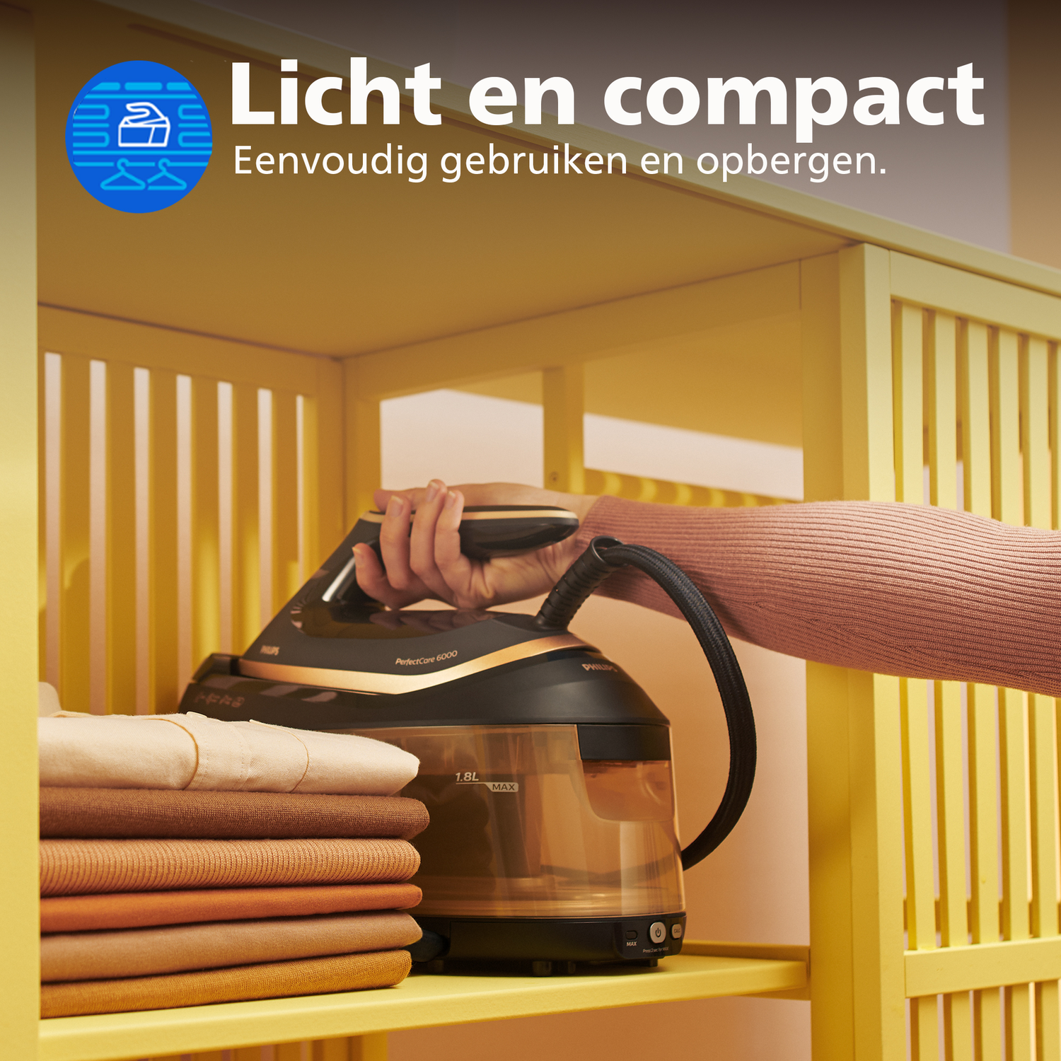 Een hand houdt een strijkijzer boven gestapelde kleding op een gele plank.