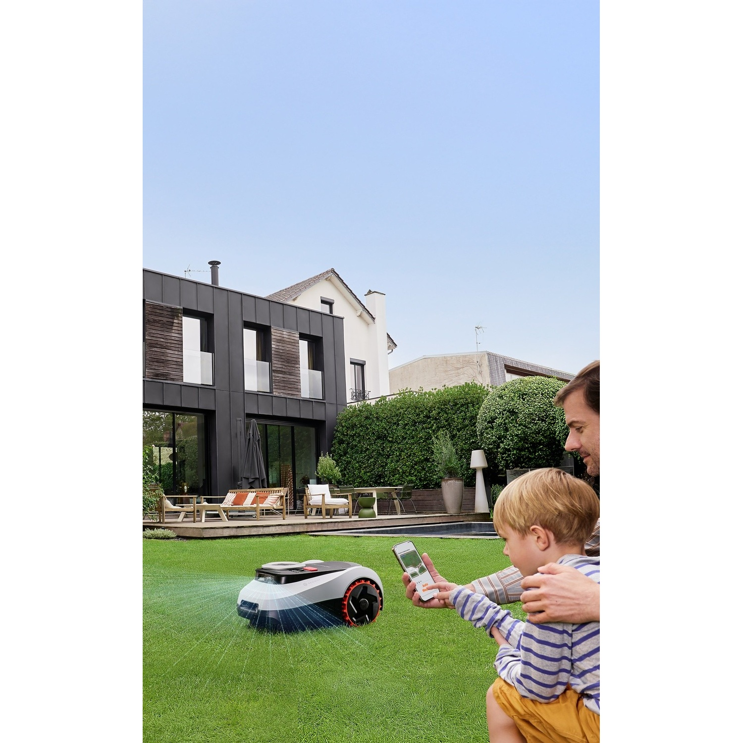 Un robot tagliaerba taglia l'erba davanti a una casa moderna; uomo e bambino lo controllano via smartphone.