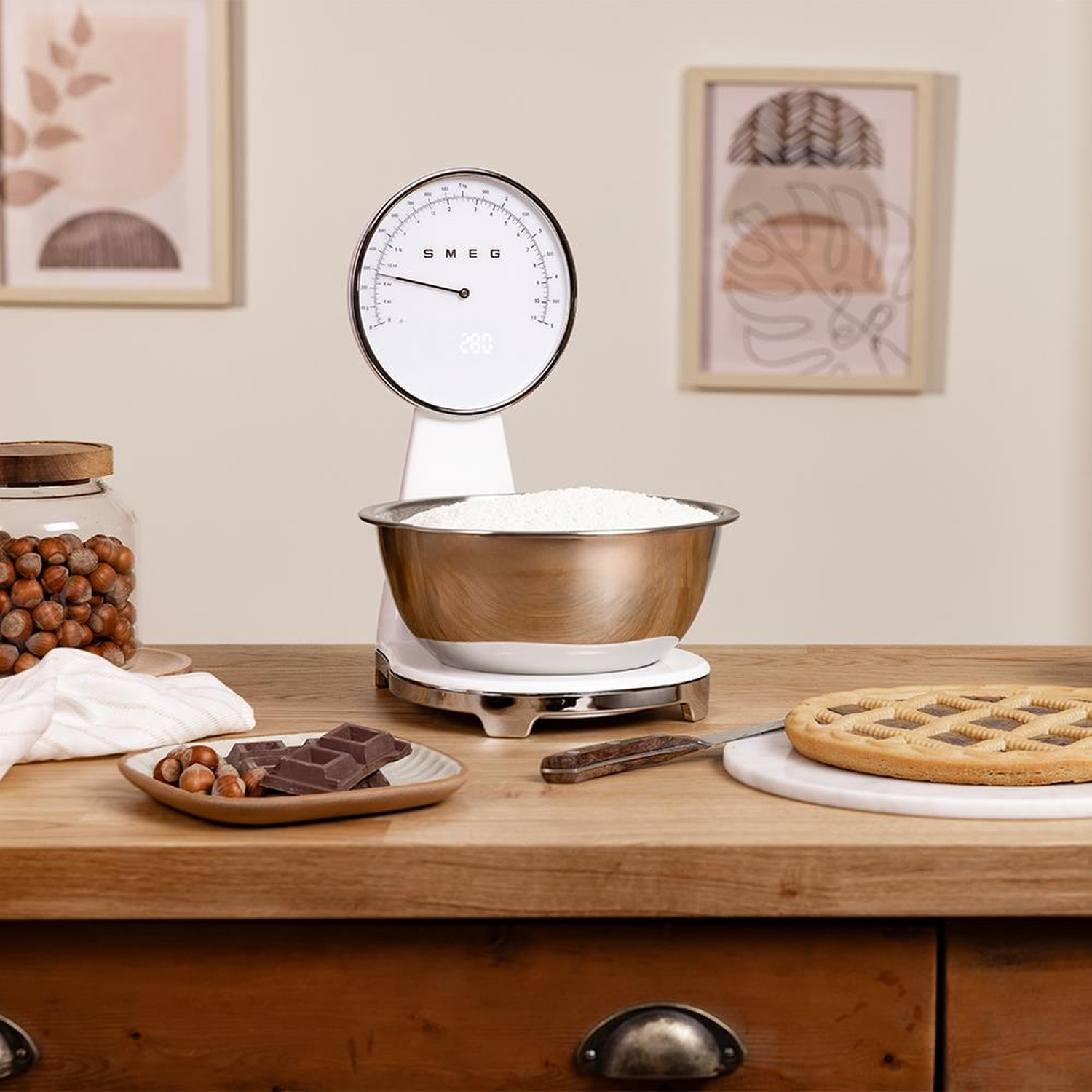 Una bilancia Smeg bianca con una ciotola di farina, cioccolato, nocciole e una crostata.