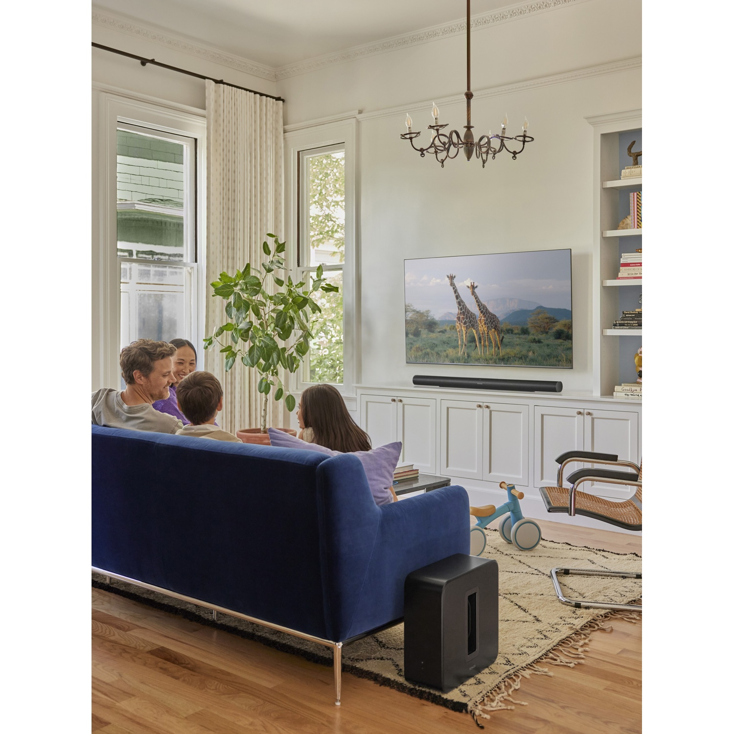 Een gezin kijkt tv in een woonkamer met een blauwe bank, boekenplanken en een tv met giraffen.