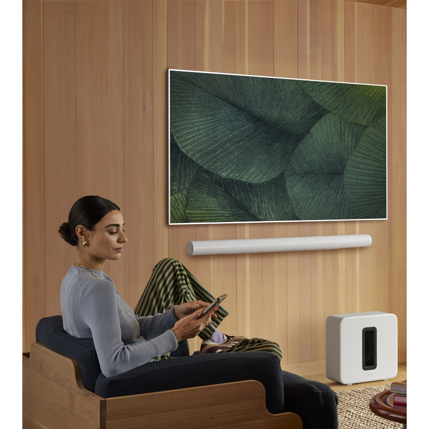 Vrouw zit in stoel, kijkt op telefoon. TV, soundbar en subwoofer op houten muur.