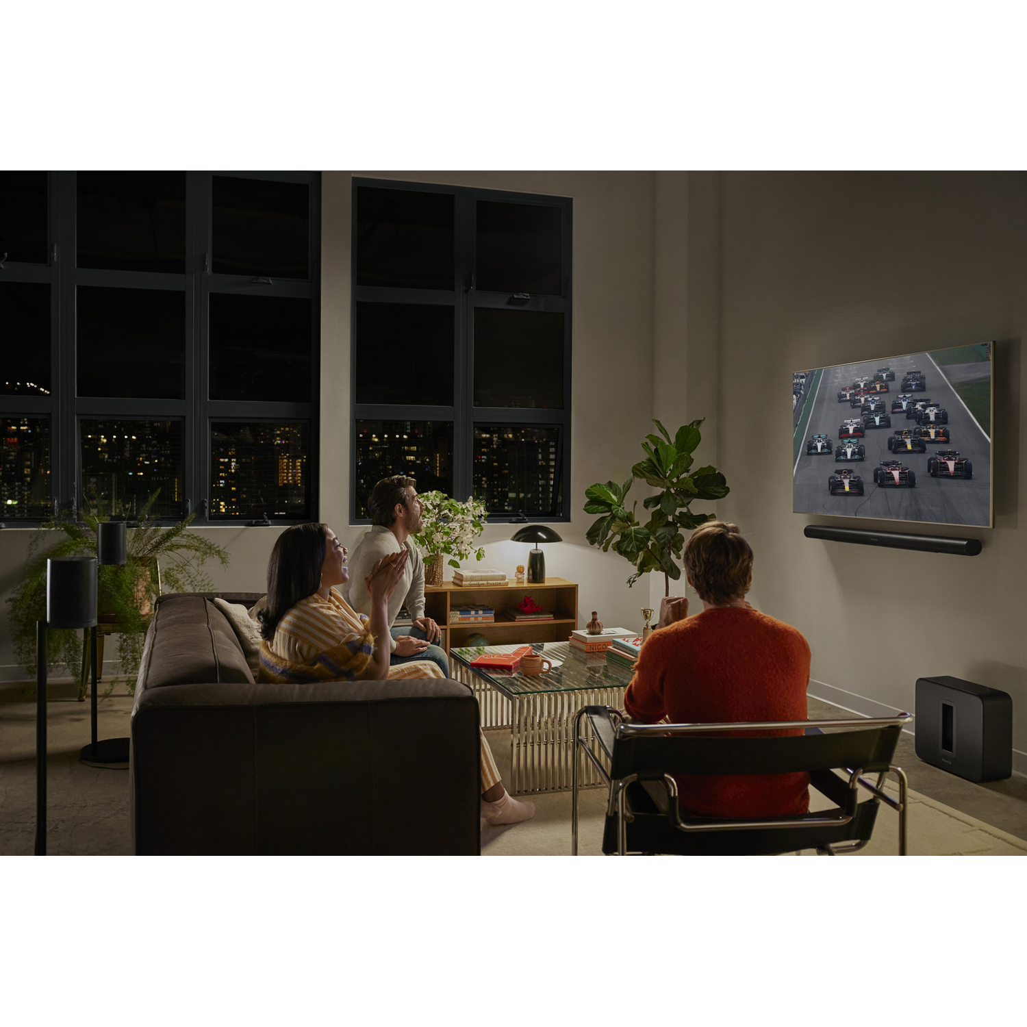 Drie mensen kijken naar een autorace op tv in een woonkamer.