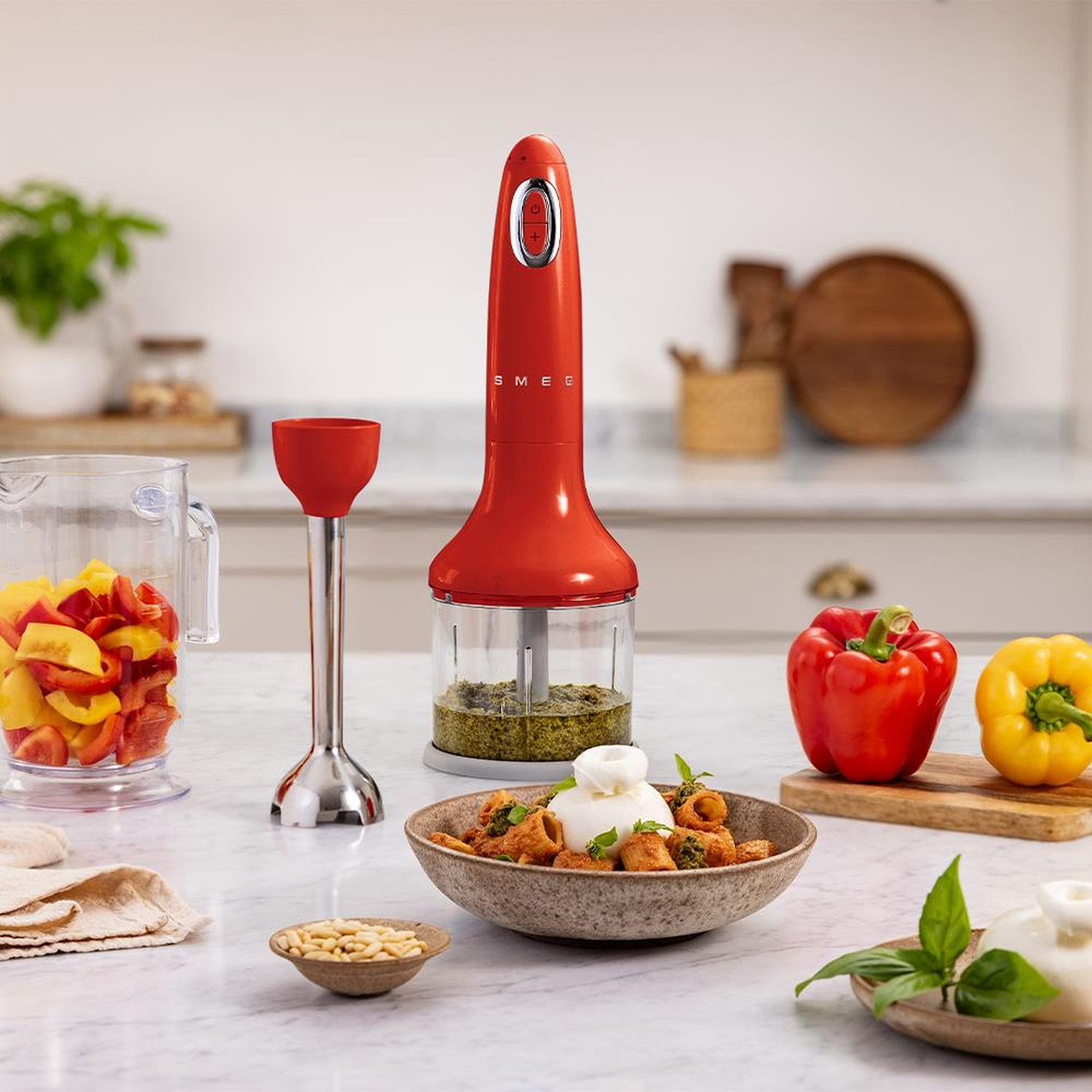 Frullatore a immersione rosso con accessori, peperoni tritati, pesto, burrata e pinoli.