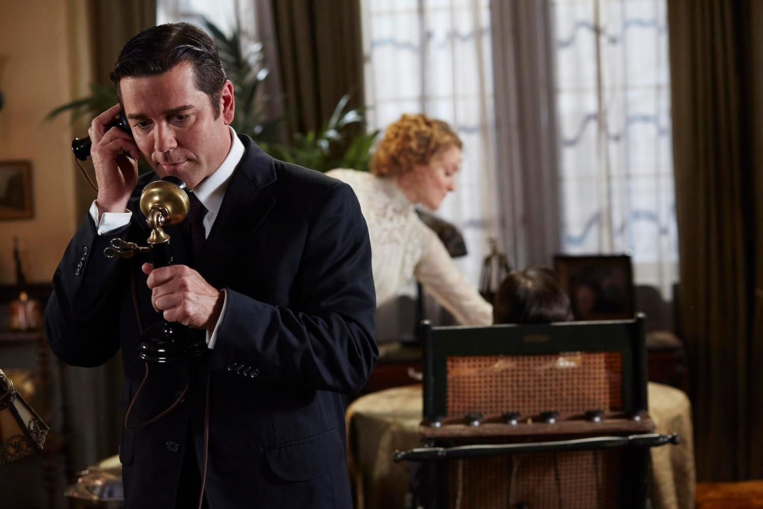 Mann im Anzug am Vintage-Telefon, Frau im Hintergrund.