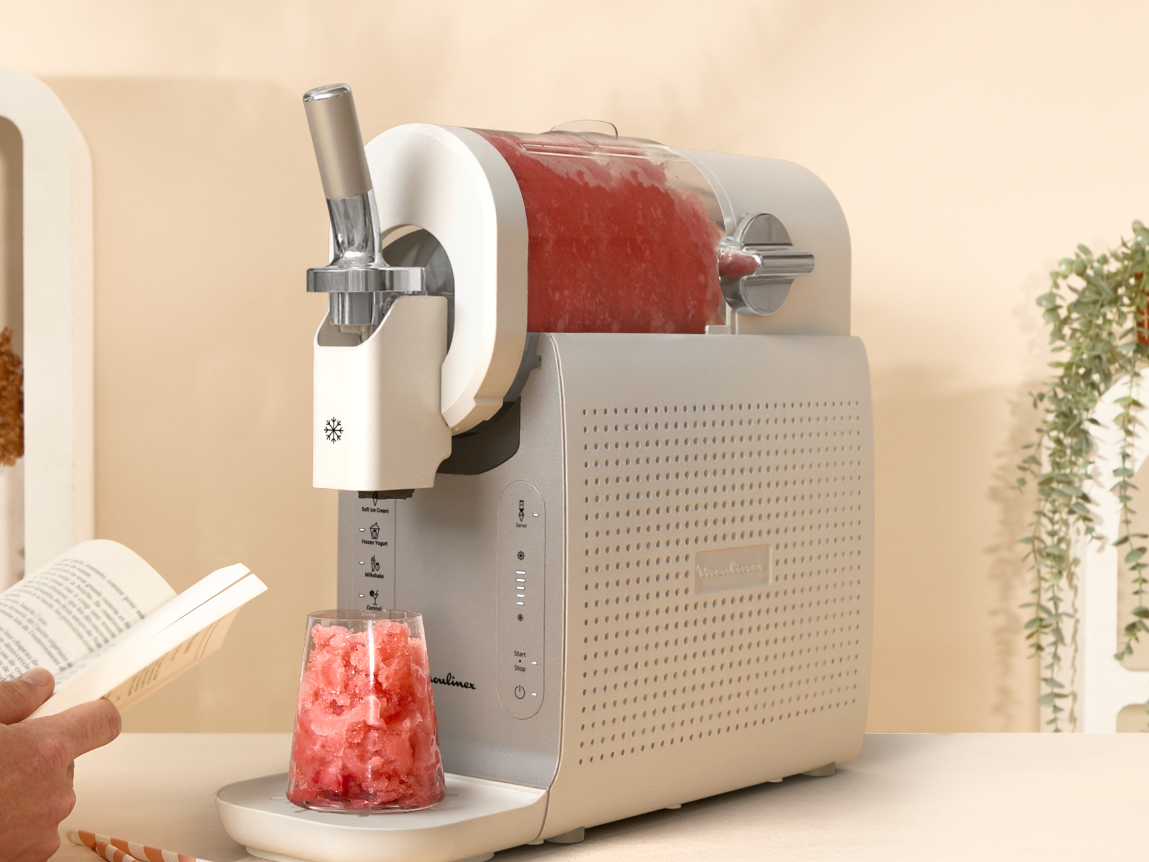 Una máquina para granizados con contenido rojo junto a un libro y un vaso de hielo picado.