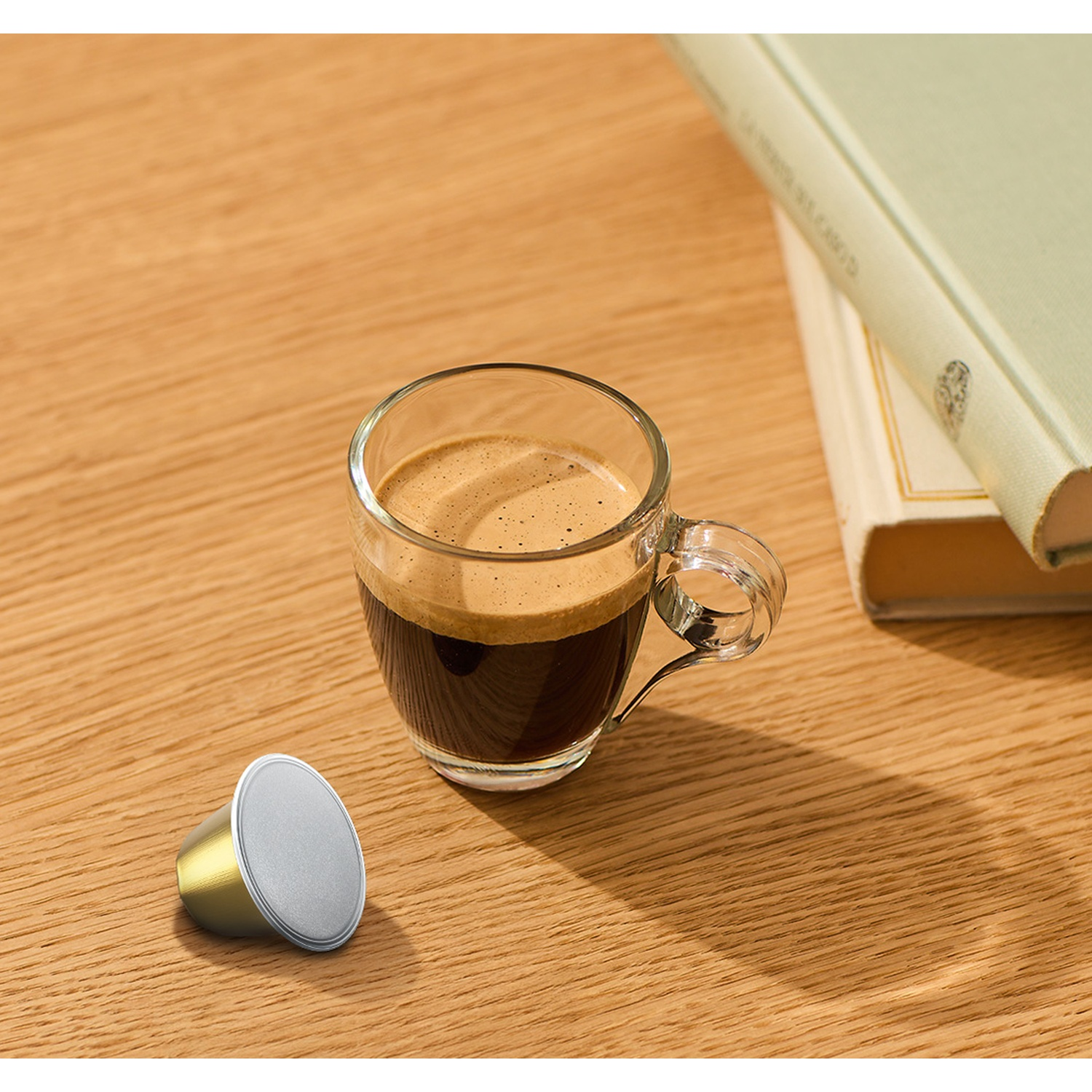 Una tazza di espresso in vetro accanto a una capsula di caffè su un tavolo di legno, con libri sullo sfondo.