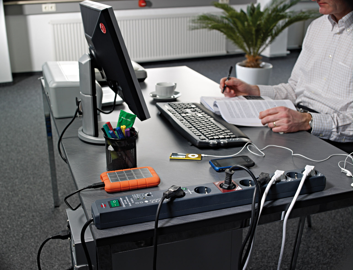 Schreibtisch mit Computer, Tastatur, Solar-Ladegerät, Steckdosenleiste und Mann, der liest.