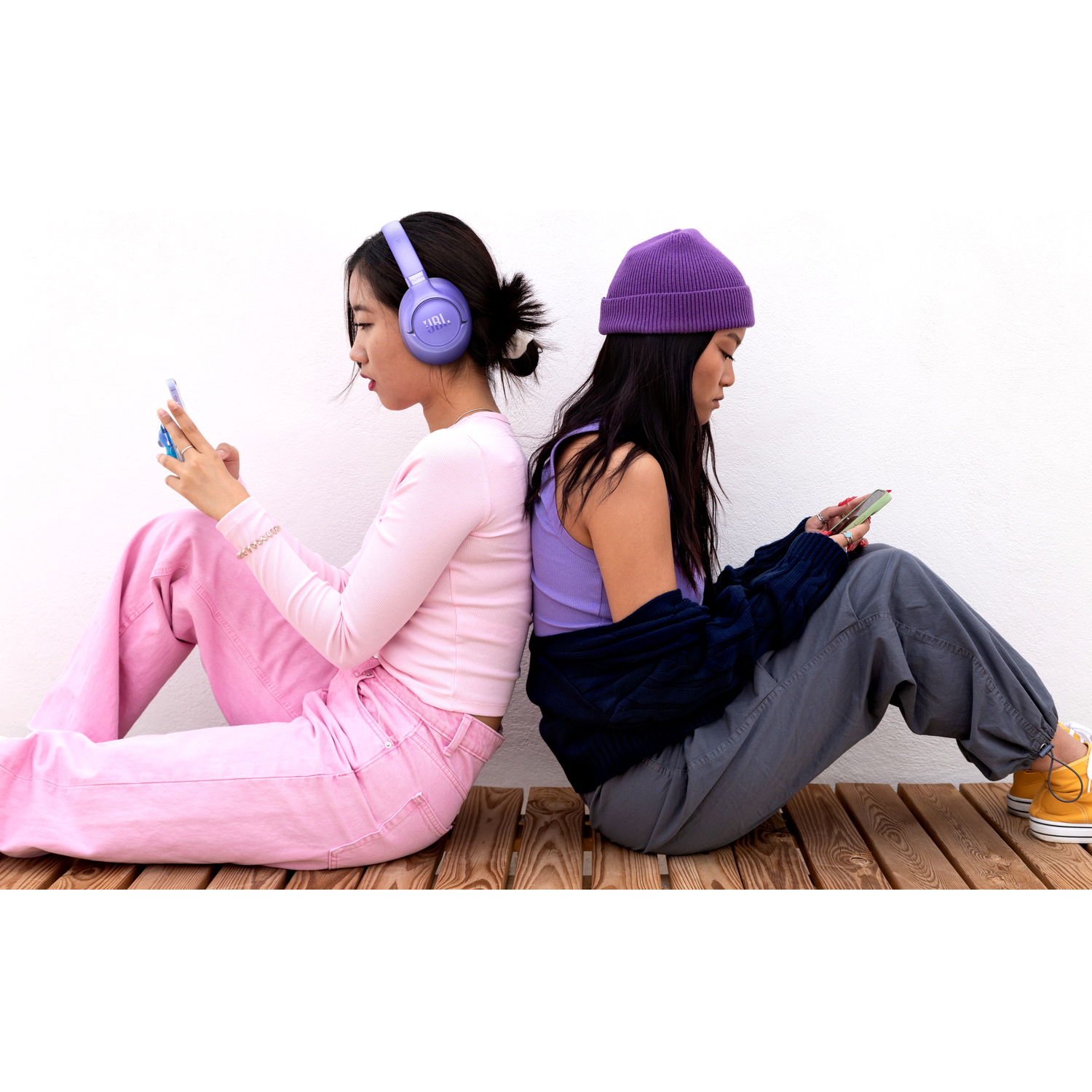 Twee jonge Aziatische vrouwen zitten rug aan rug op houten planken, elk verdiept in hun telefoon.
