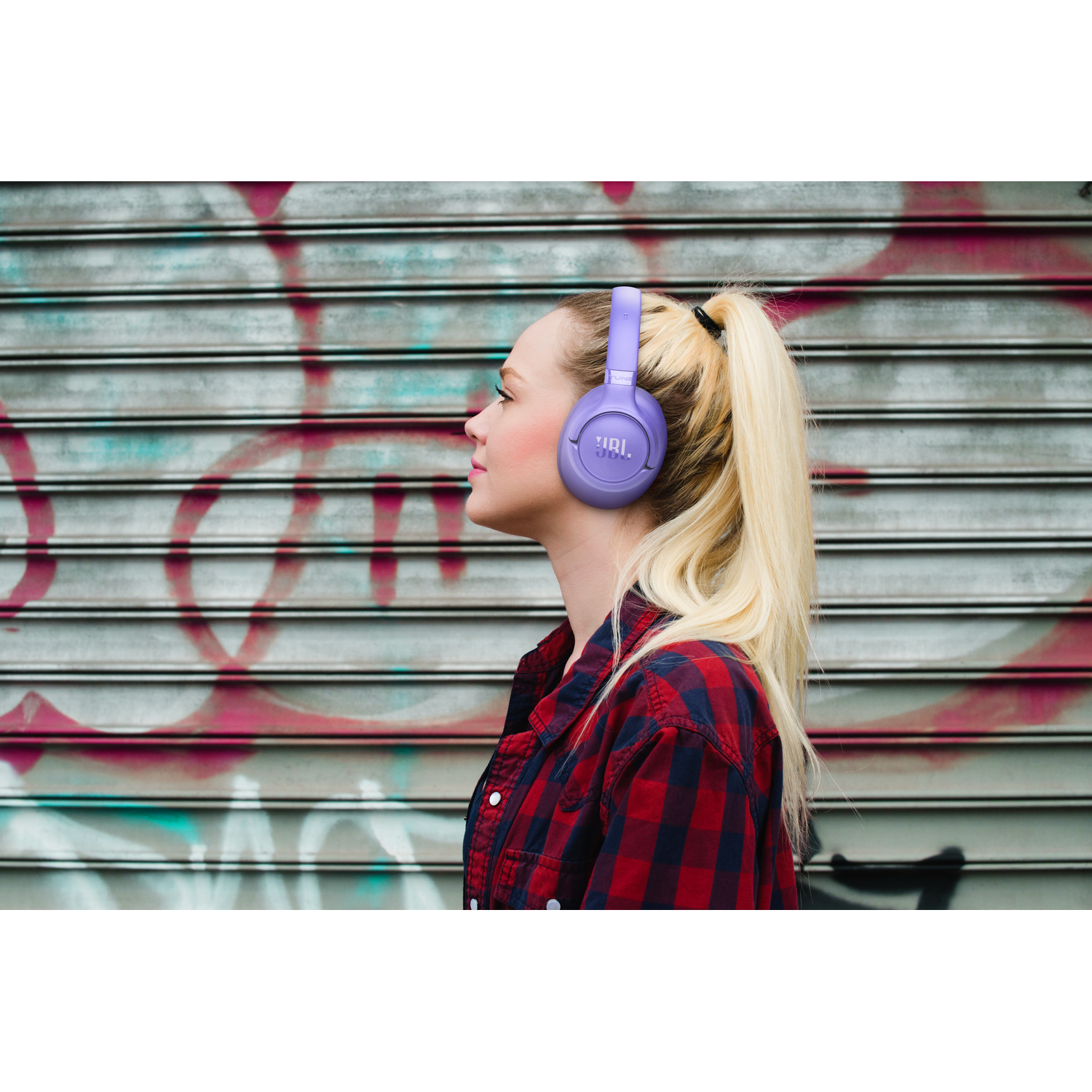 Vrouw met blonde paardenstaart draagt paarse koptelefoon, profiel, tegen graffiti muur.