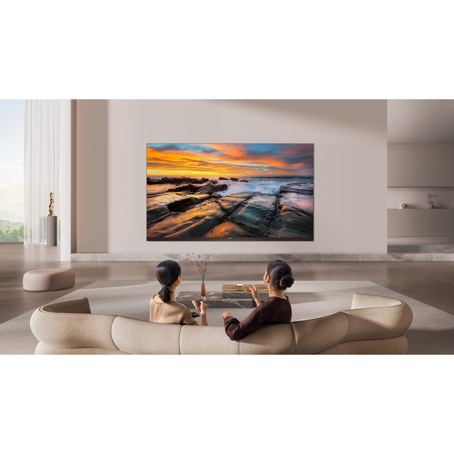Twee vrouwen zitten op een bank en kijken naar een grote tv met zonsondergang op rotsen.