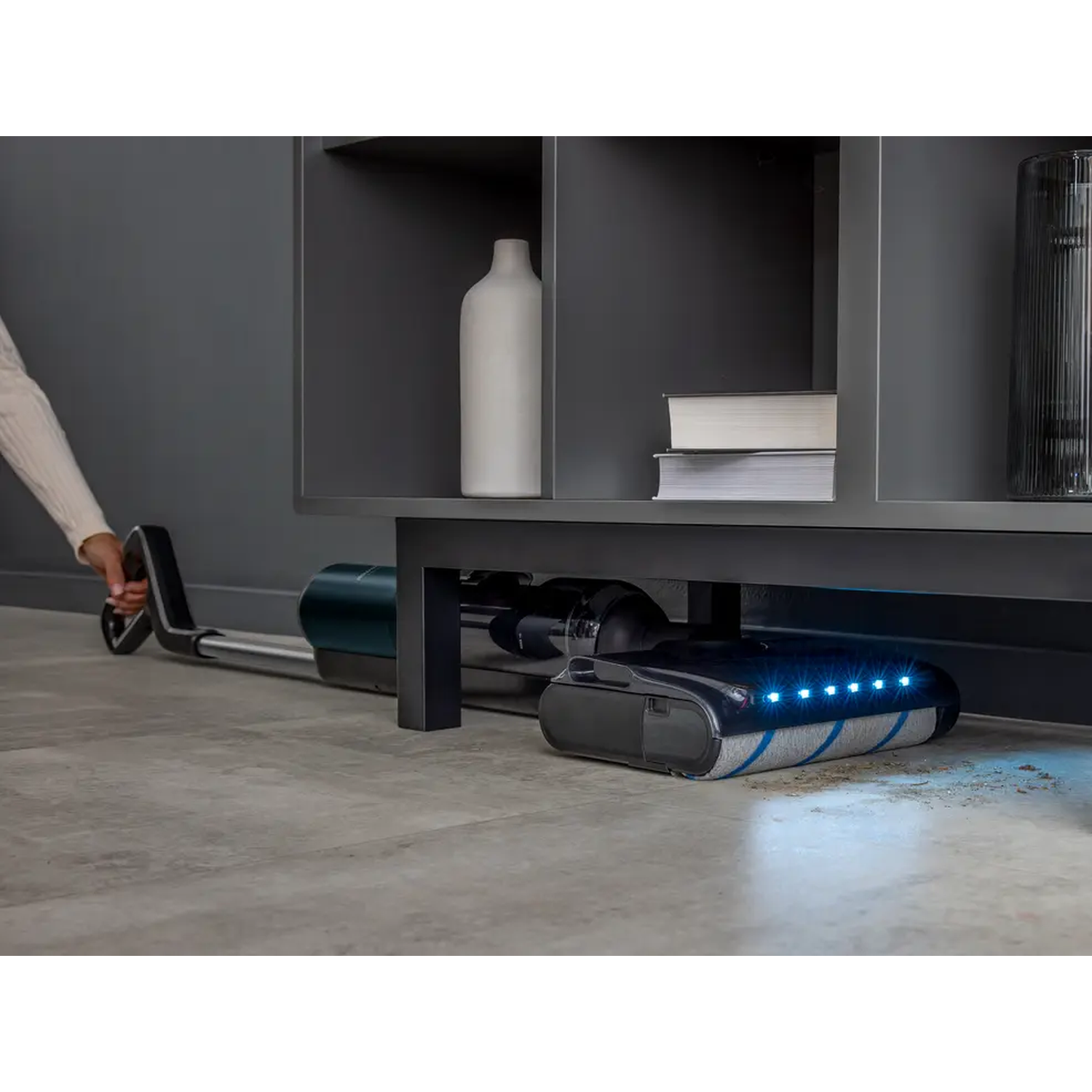 A handheld vacuum cleaner cleans a floor under a shelf with books and a vase.
