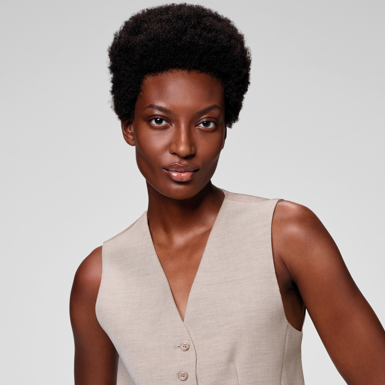 Een vrouw met donkere huid en kort afro haar draagt een beige vest.