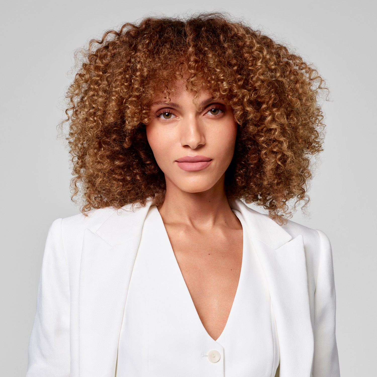 Een vrouw met krullend bruin haar draagt een witte blazer en kijkt vooruit.