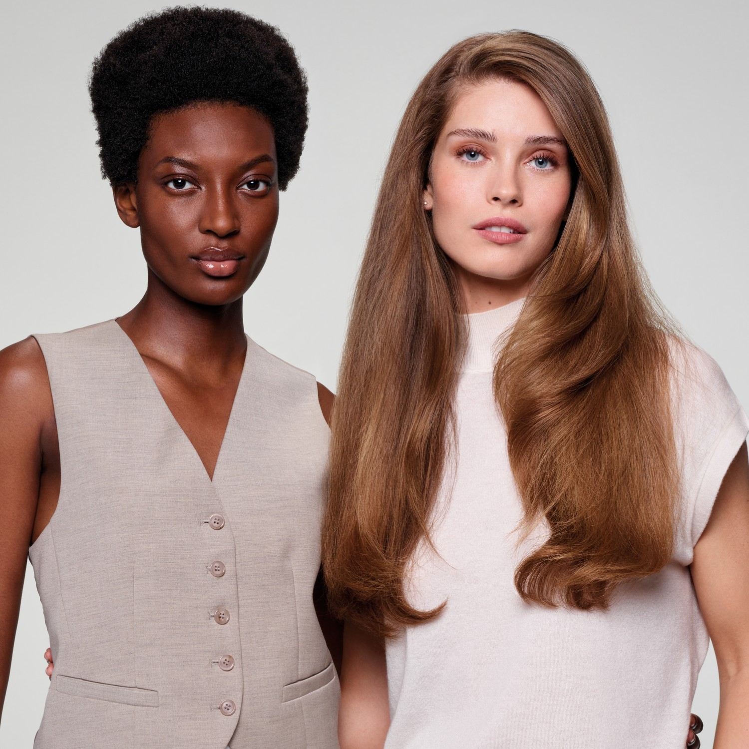 Twee vrouwen staan naast elkaar. Eén heeft een donkere huid en kort afrohaar. De ander heeft een lichte huid en lang, bruin haar.