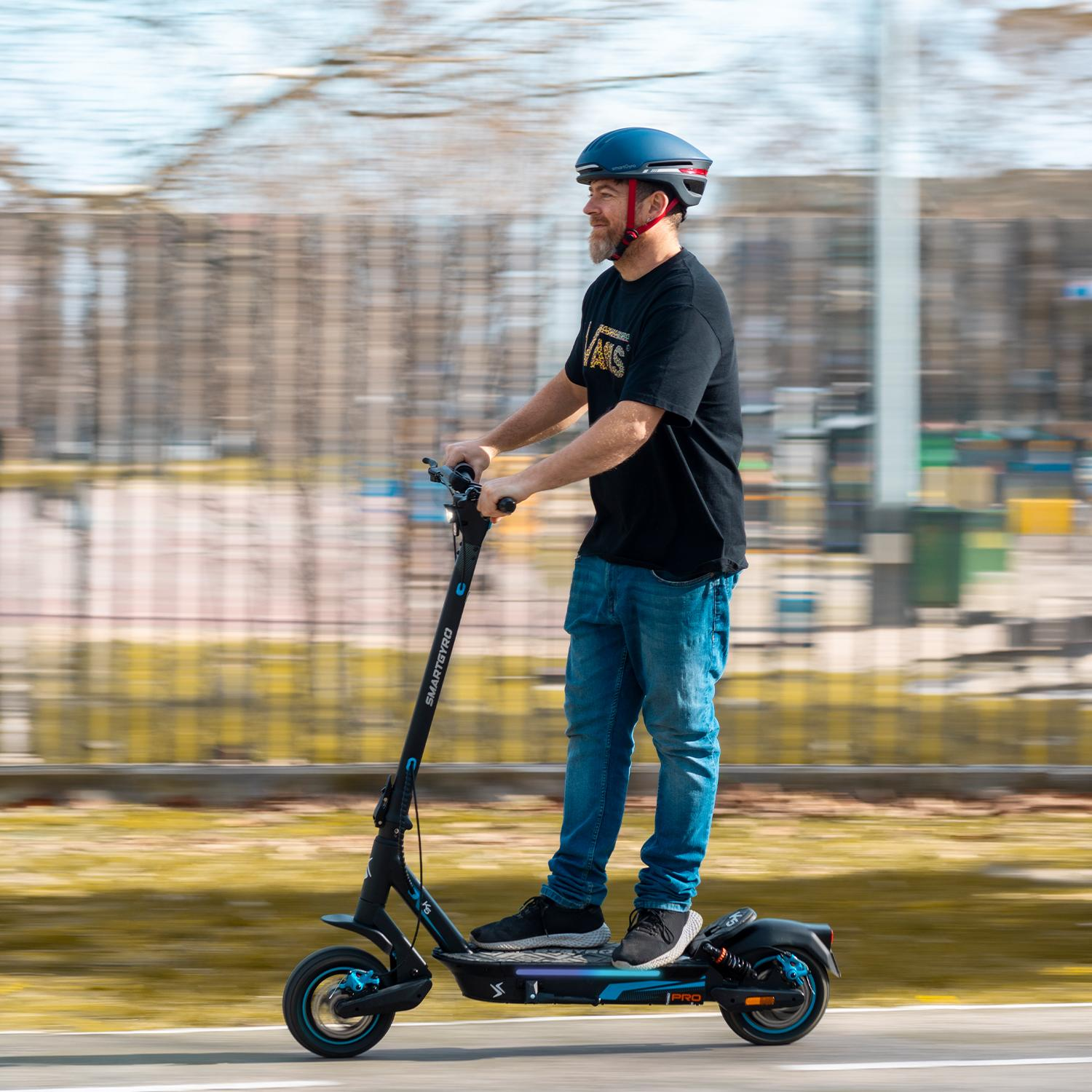 Hombre monta patinete eléctrico, casco azul, camiseta negra, vaqueros en calle.