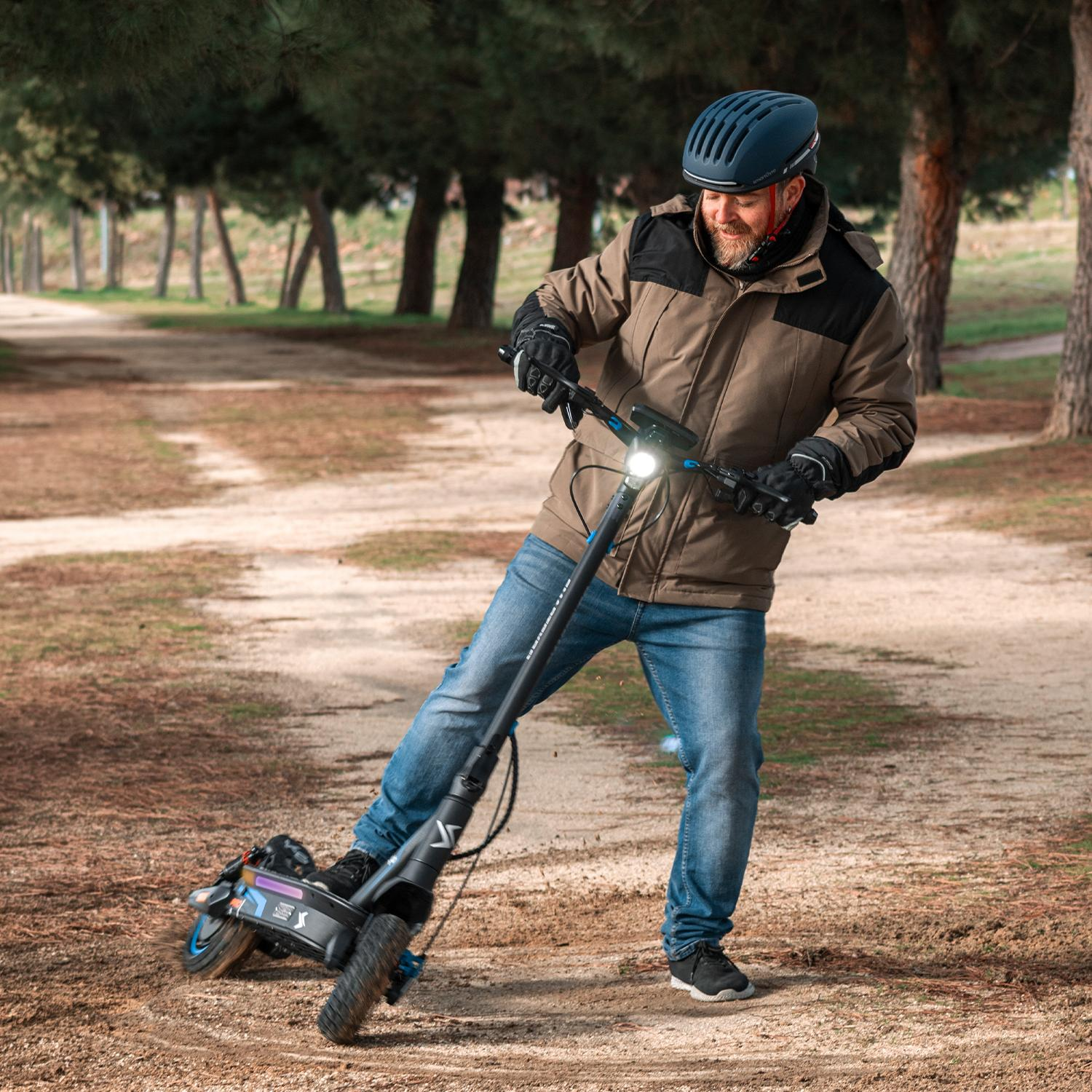 Hombre con casco monta patinete eléctrico en camino de tierra, rodeado de árboles.