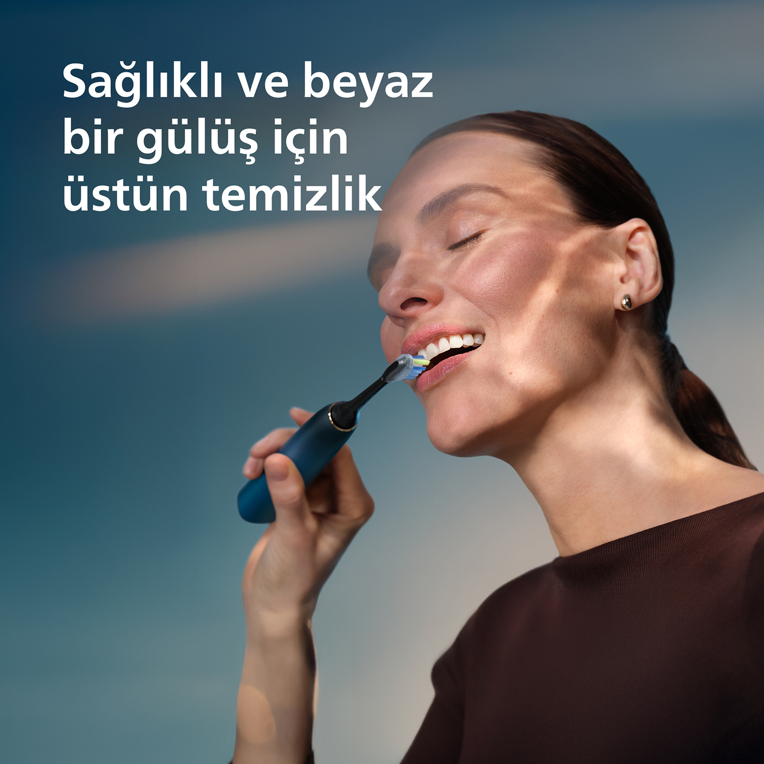 Kadın mavi elektrikli diş fırçasıyla mavi arka plana karşı dişlerini fırçalıyor.