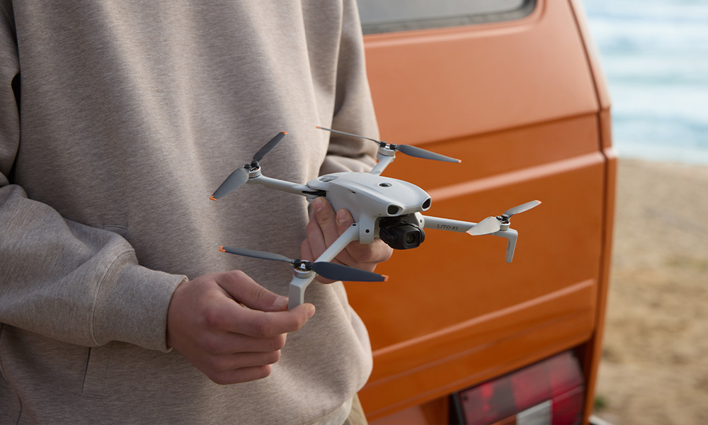 Osoba trzyma biały dron przed pomarańczowym kamperem na plaży.