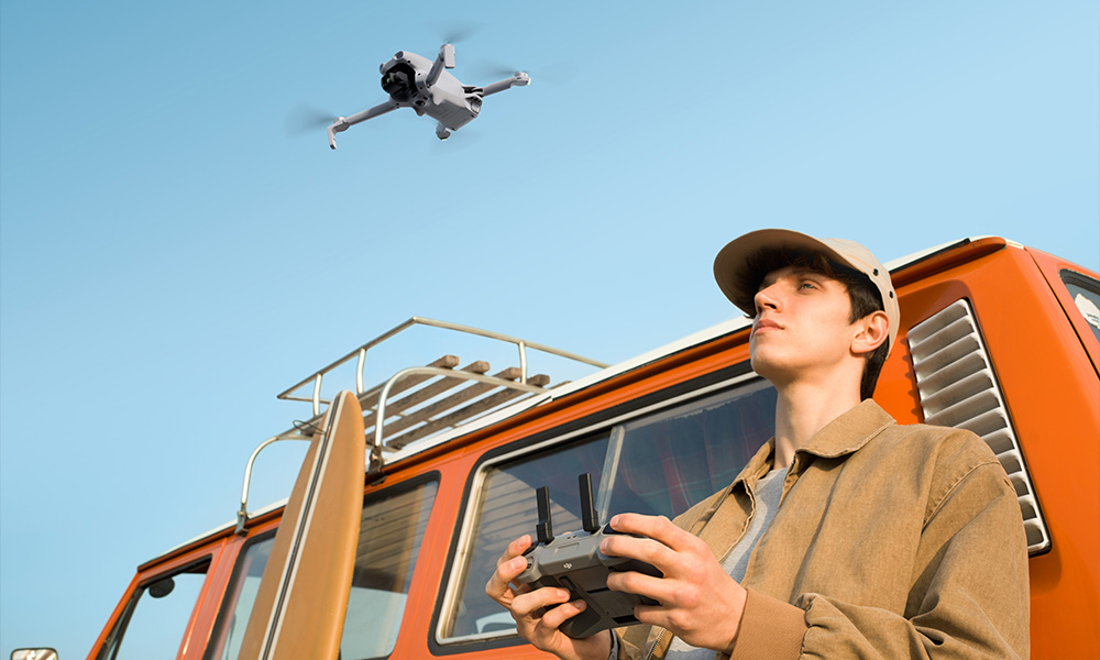 Mężczyzna steruje dronem lecącym nad kamperem z deską surfingową.