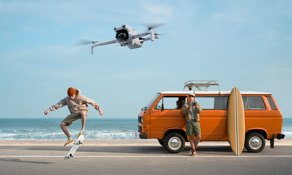 Skejtboarder skacze przed pomarańczowym vanem na plaży, leci dron.