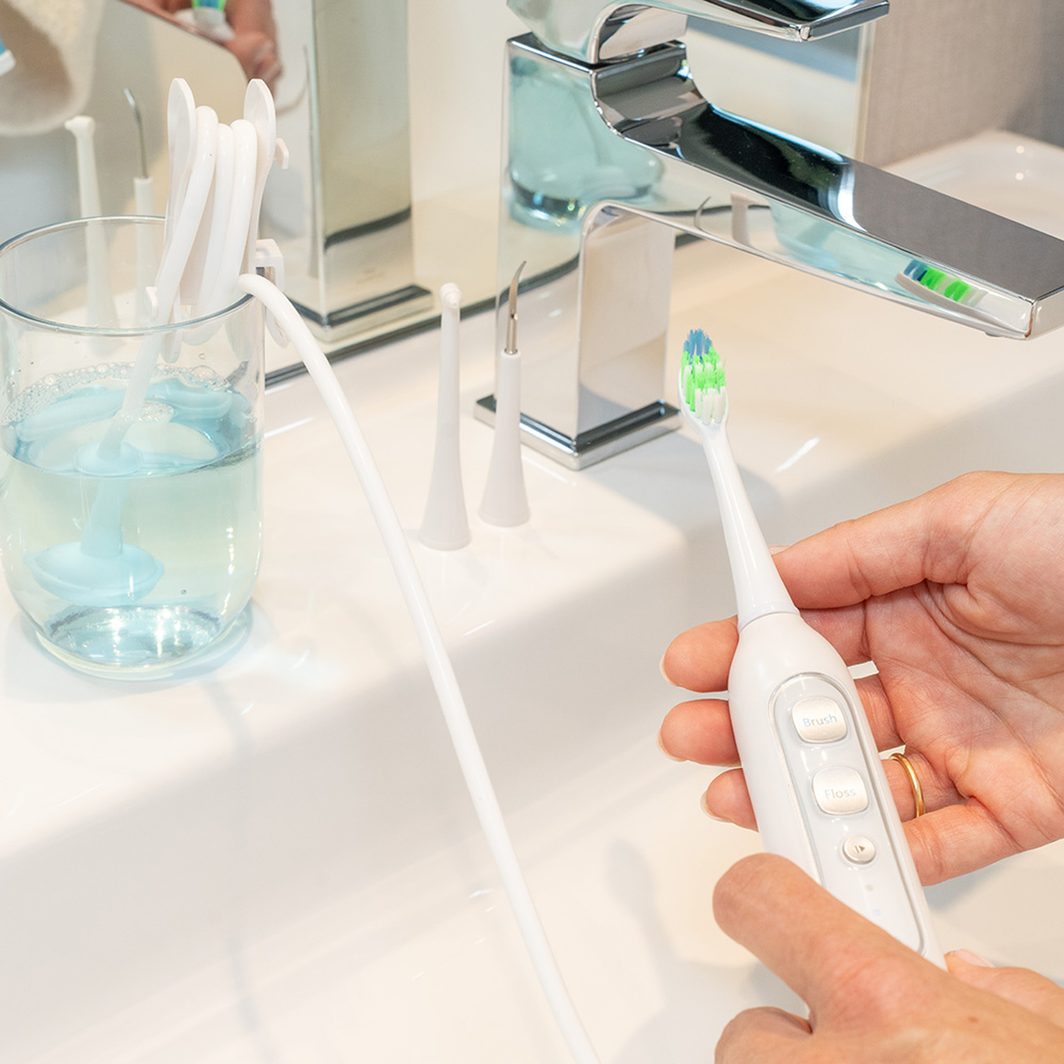 A person holds a white electric toothbrush with blue and green bristles near a sink.