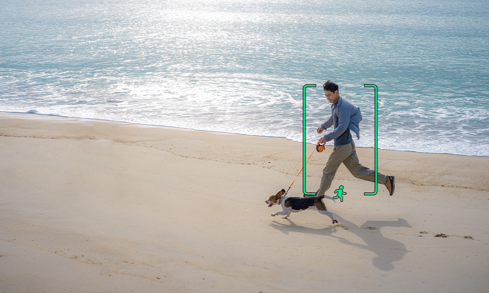 Mężczyzna w szarej kurtce i beżowych spodniach biegnie z beaglem na plaży przy niebieskiej wodzie.