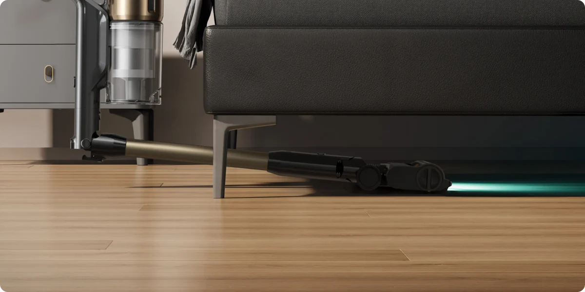 Ein Staubsauger mit grünem Licht reinigt unter einem schwarzen Sofa auf Holzboden.