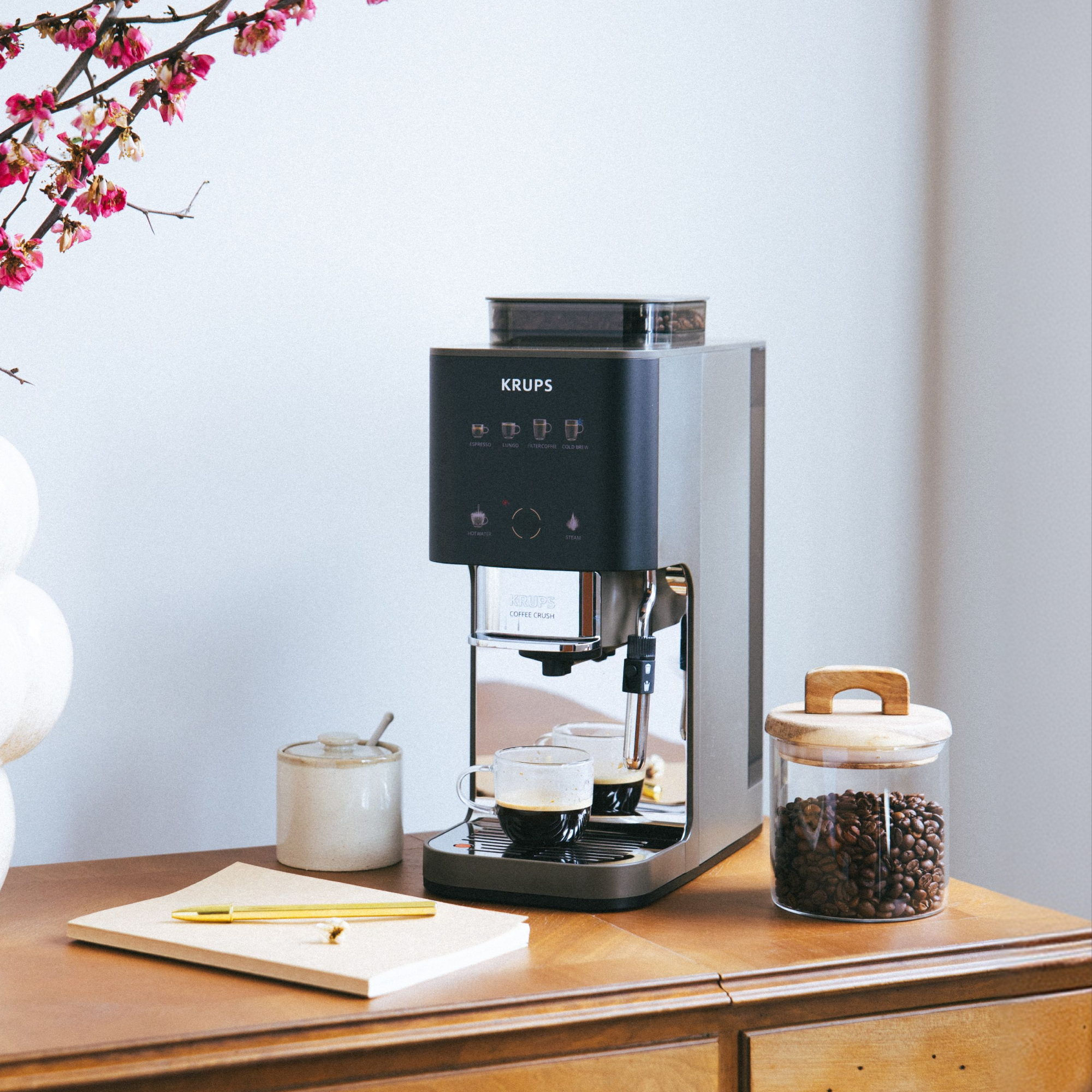 Cafetera Krups con dos tazas de espresso, tarro de granos y libreta.