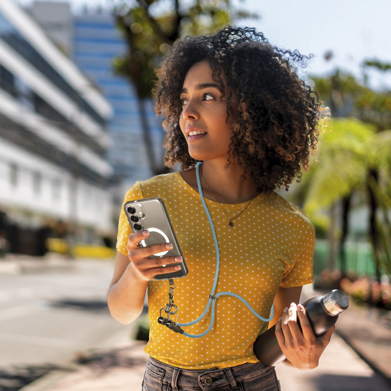 Donna con capelli ricci, maglia gialla a pois, tiene telefono e borraccia.