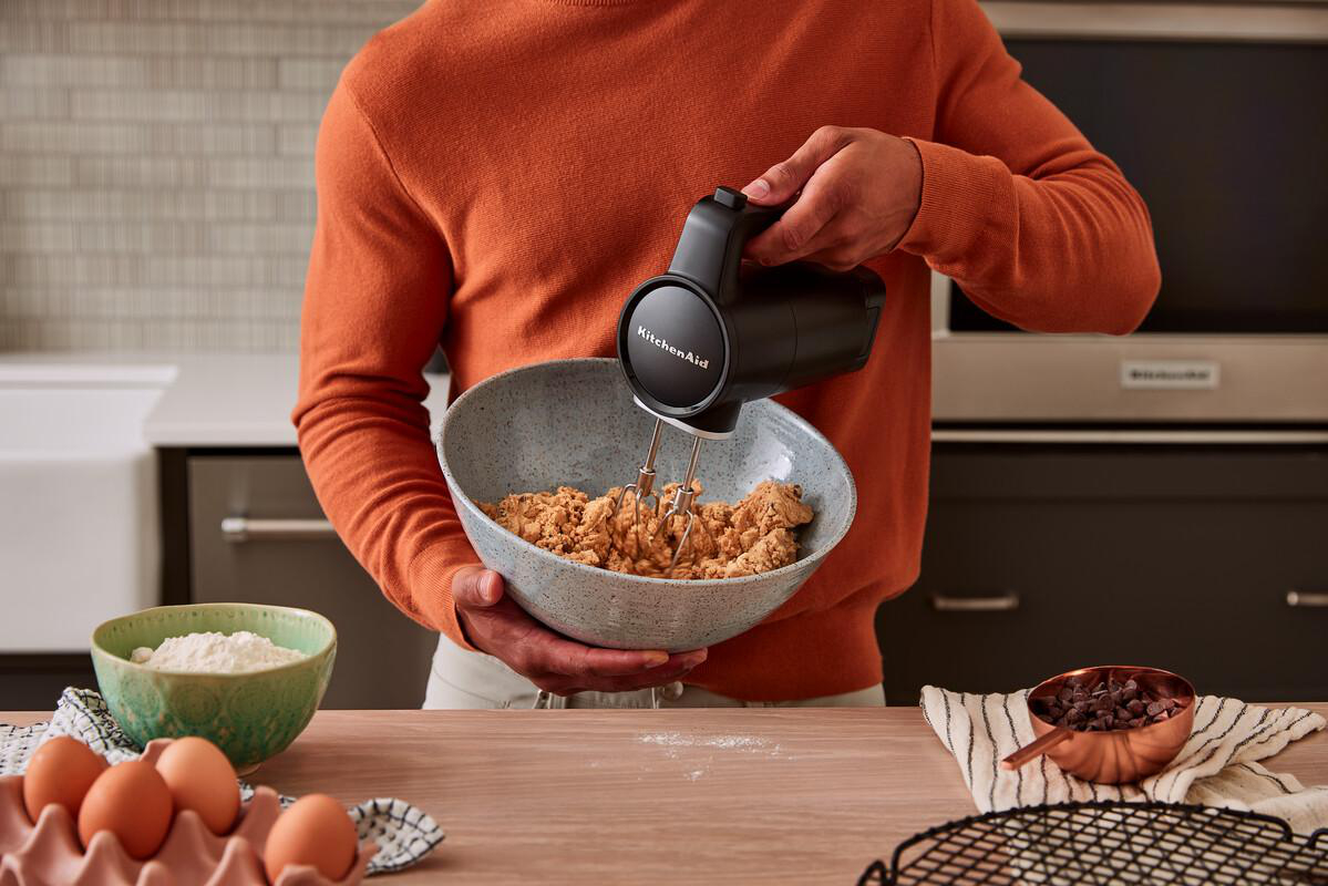 Eine Person in einem orangefarbenen Pullover mischt mit einem schwarzen Handmixer in einer Schüssel.