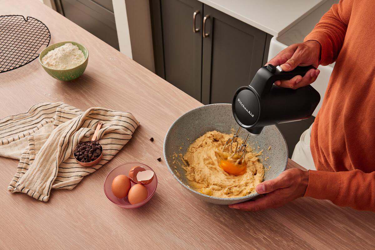Eine Person mixt Teig mit einem schwarzen Handmixer in einer Schüssel. Zutaten wie Eier und Mehl sind auf dem Tisch.