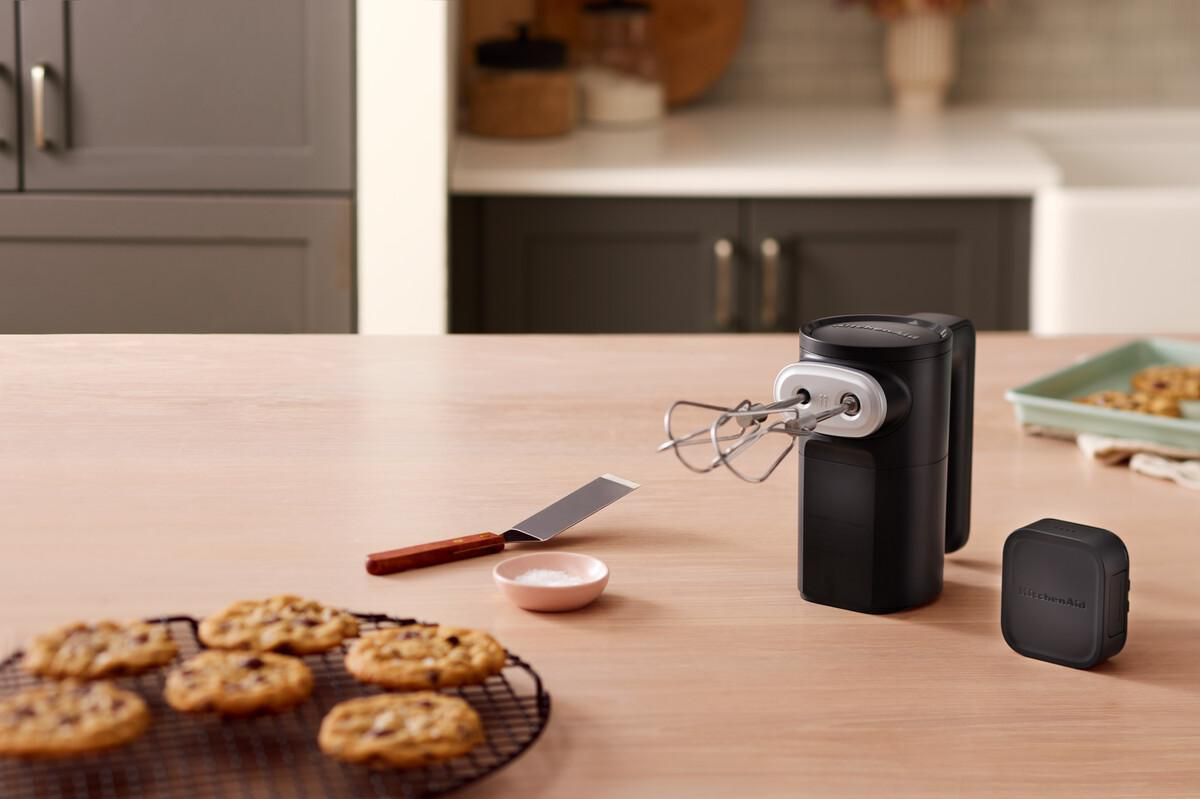 Ein schwarzer Handmixer mit Schneebesen neben Keksen auf einem Holztisch.