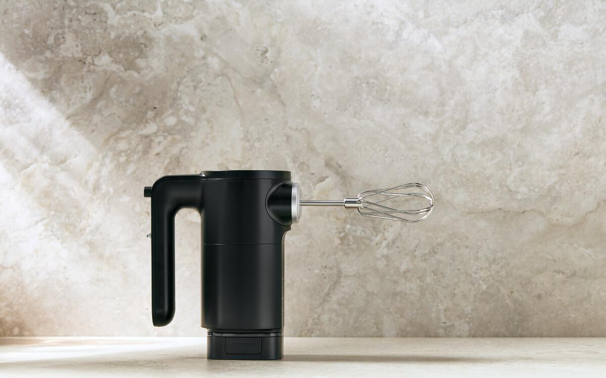 Schwarzer Handmixer mit Quirl auf einer Steinplatte vor hellem Hintergrund.