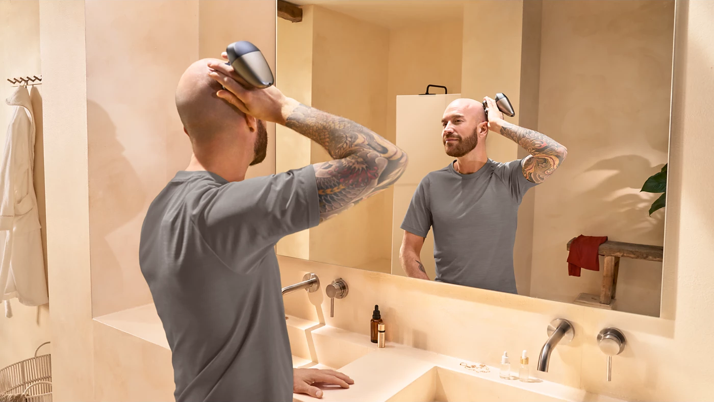 Ein Mann mit Tattoos rasiert sich den Kopf vor einem Spiegel im Badezimmer.