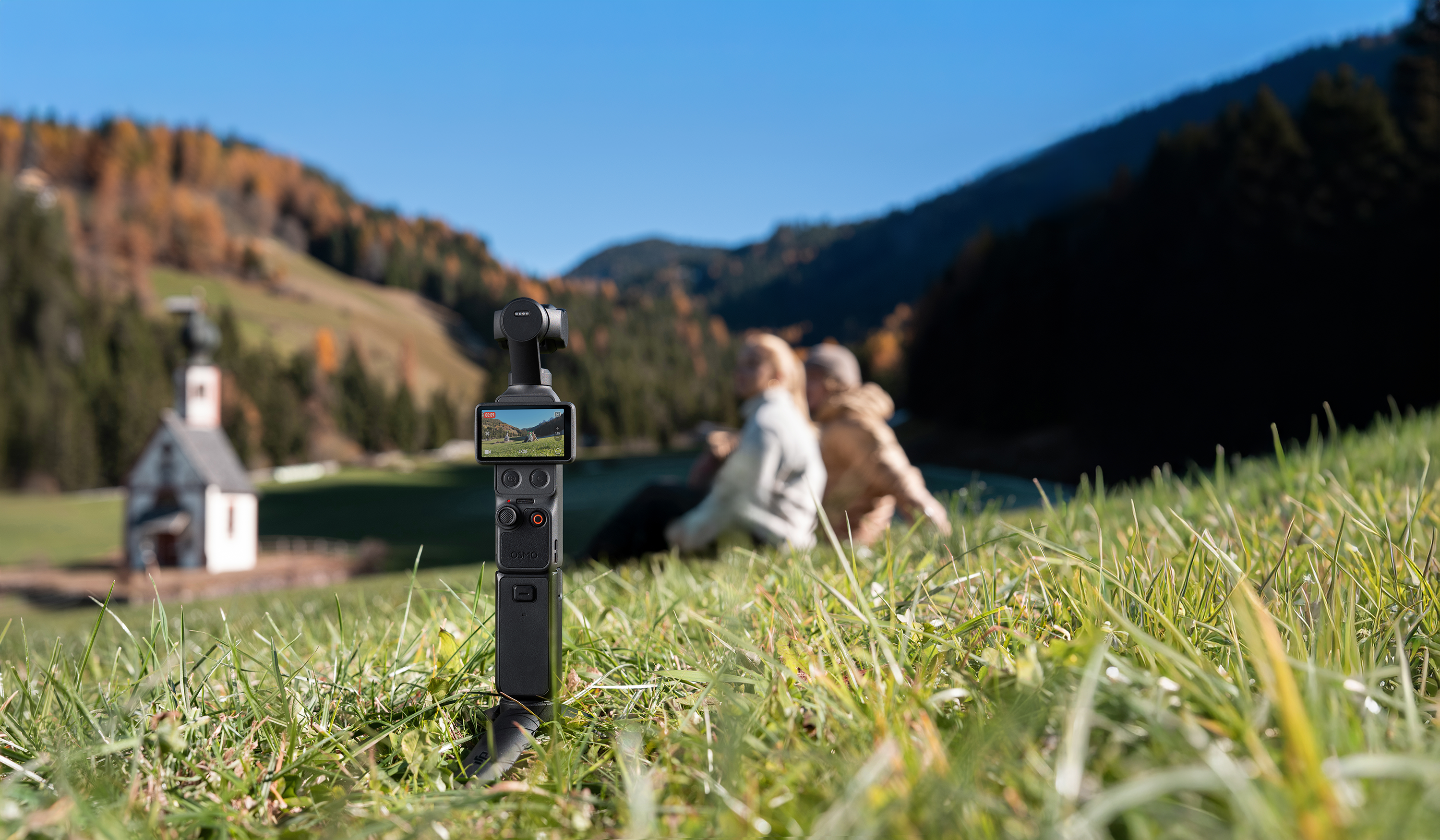 Egy gimbal stabilizátor áll az előtérben zöld füvön. A háttérben két ember, egy templom és egy domb látható.
