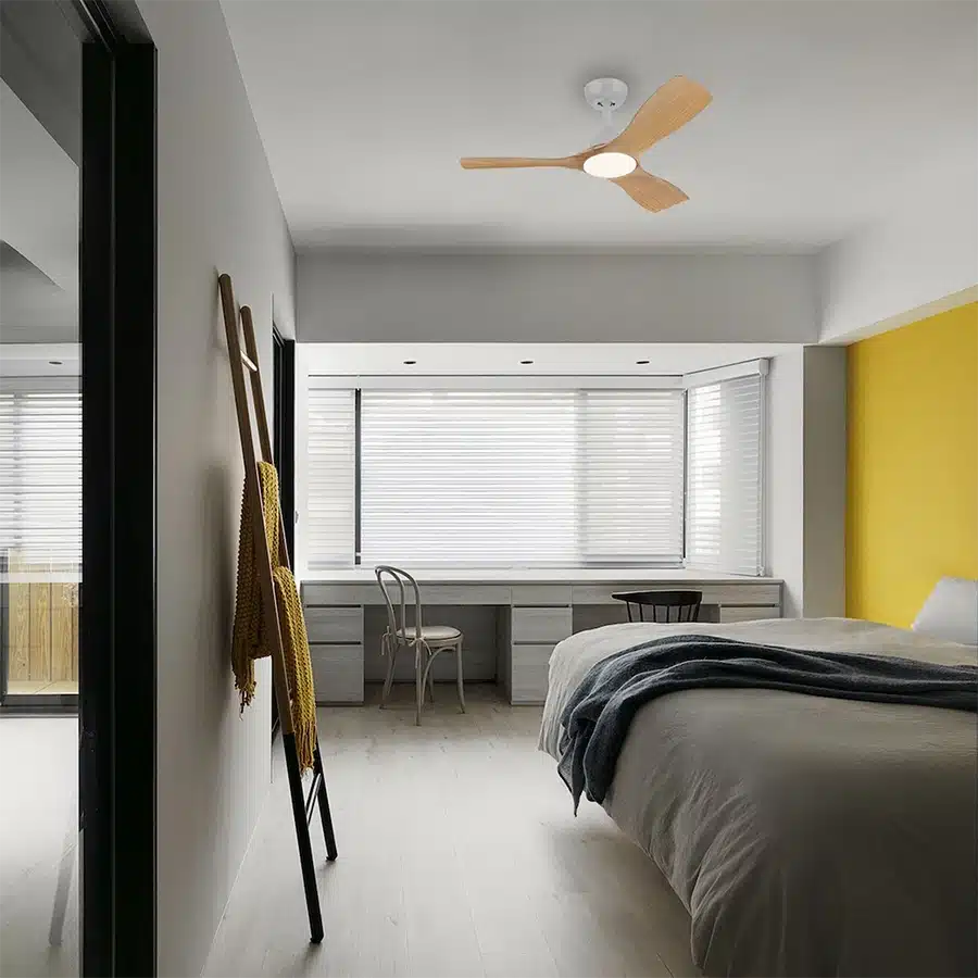 Un dormitorio con cama, escritorio, silla y ventilador de techo de madera. Pared amarilla.